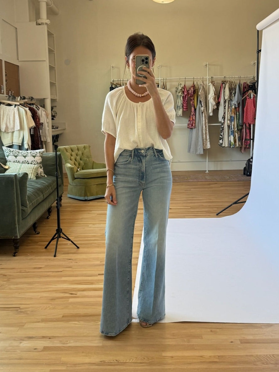Person taking a mirror selfie wearing a white top and blue jeans in a room with a white backdrop and clothing racks.