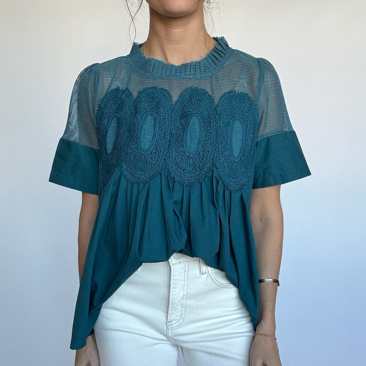 Person wearing a teal blouse with lace details and white pants against a plain background